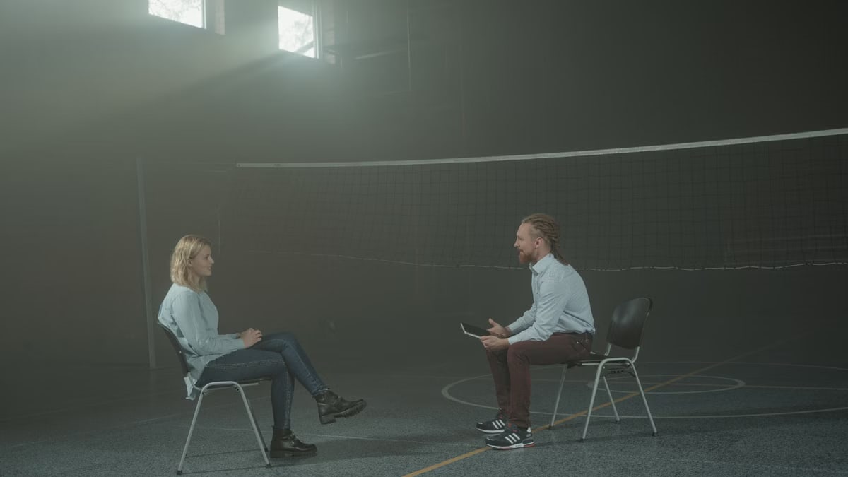A man and a woman engage in a therapy session in a smoky indoor setting, sitting apart in a large room.